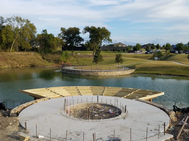 Wide view of a community amenity site under construction, showing a curved dock deck being framed beside a poured concrete plaza and fire pit.