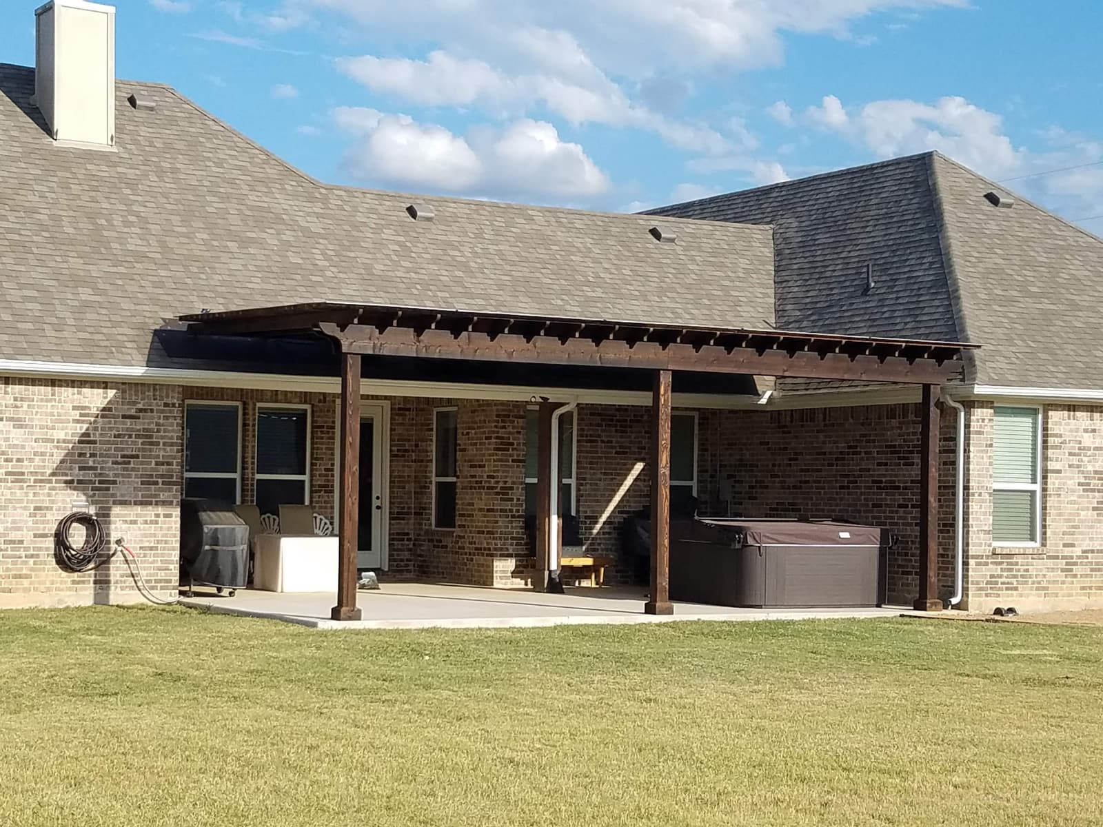 Long attached pergola with dark-stained rafters running the width of a brick home, shading a concrete patio, grill and hot tub.