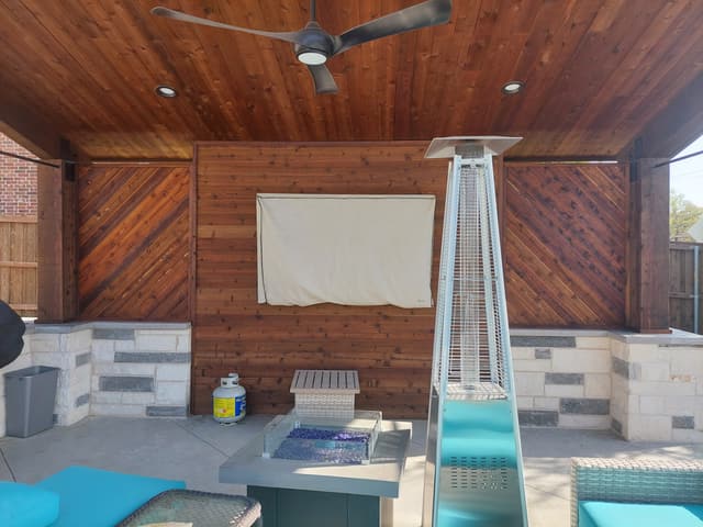 Interior of a cedar cabana with a stained plank ceiling, ceiling fan, recessed lights, a covered television and a stainless fire table.