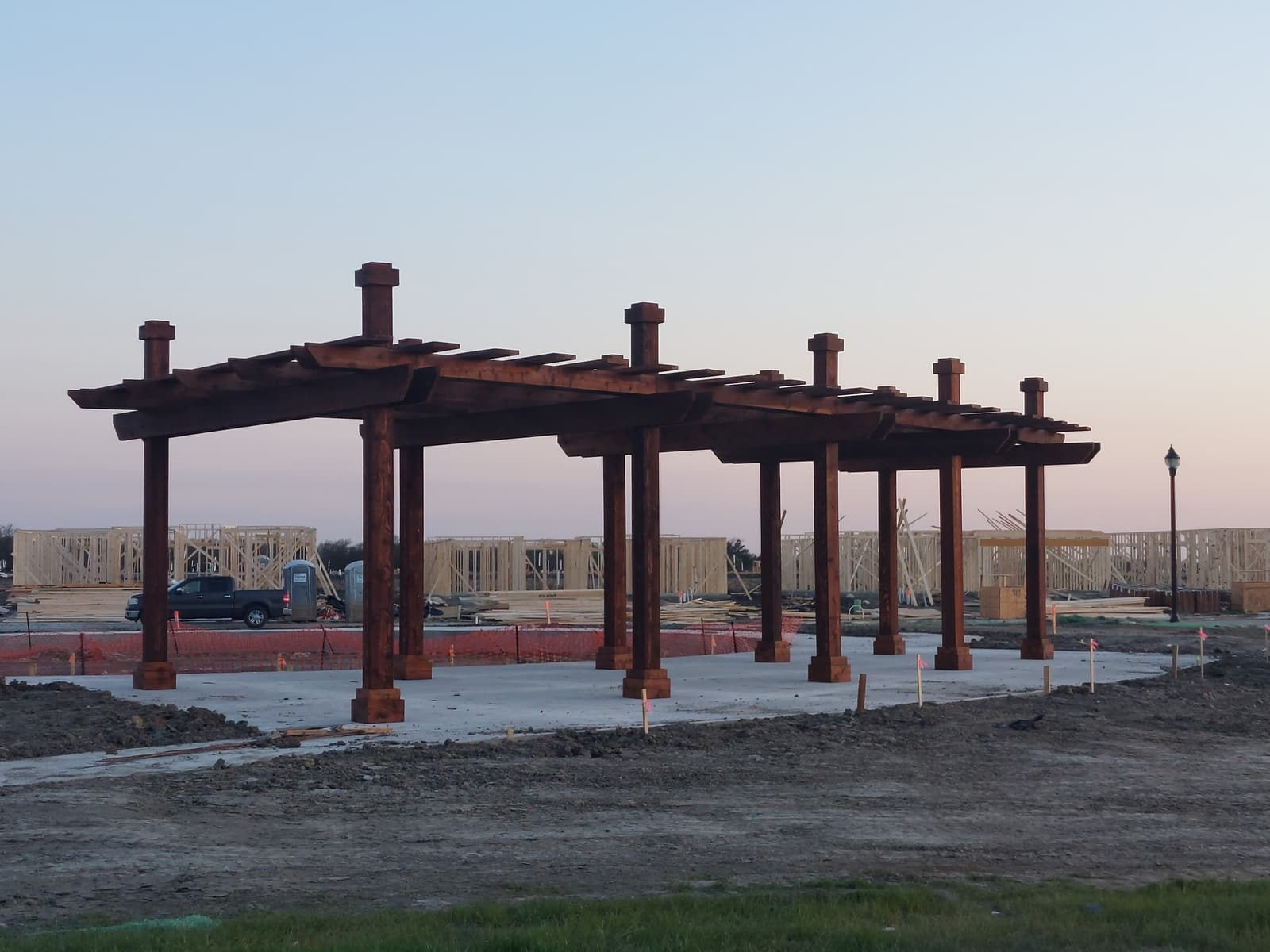 Large freestanding commercial pergola with heavy stained timber posts and beams on a concrete pad, photographed at dusk in a new development.