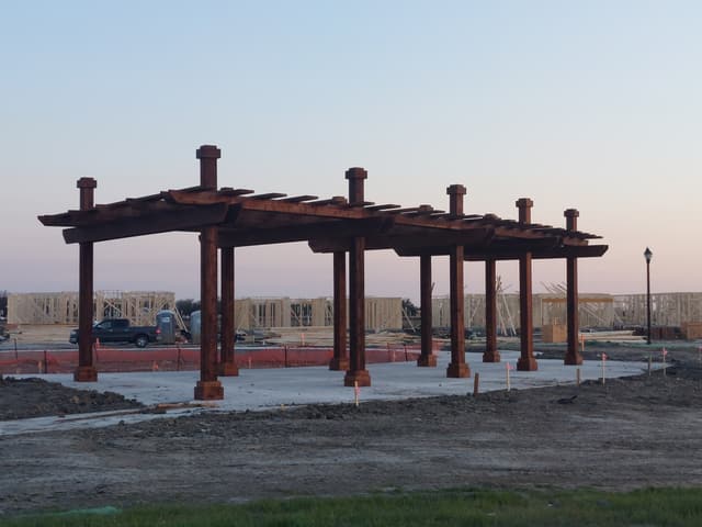 Large freestanding commercial pergola with heavy stained timber posts and beams on a concrete pad, photographed at dusk in a new development.