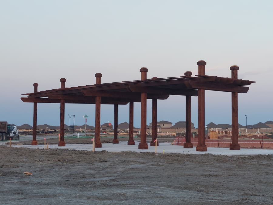 Side view at dusk of a long commercial timber pergola on a concrete pad, with a residential development under construction behind it.