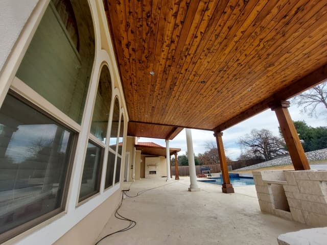 Covered patio with a richly stained cedar plank ceiling and cedar posts, running alongside a stucco home with tall arched windows.