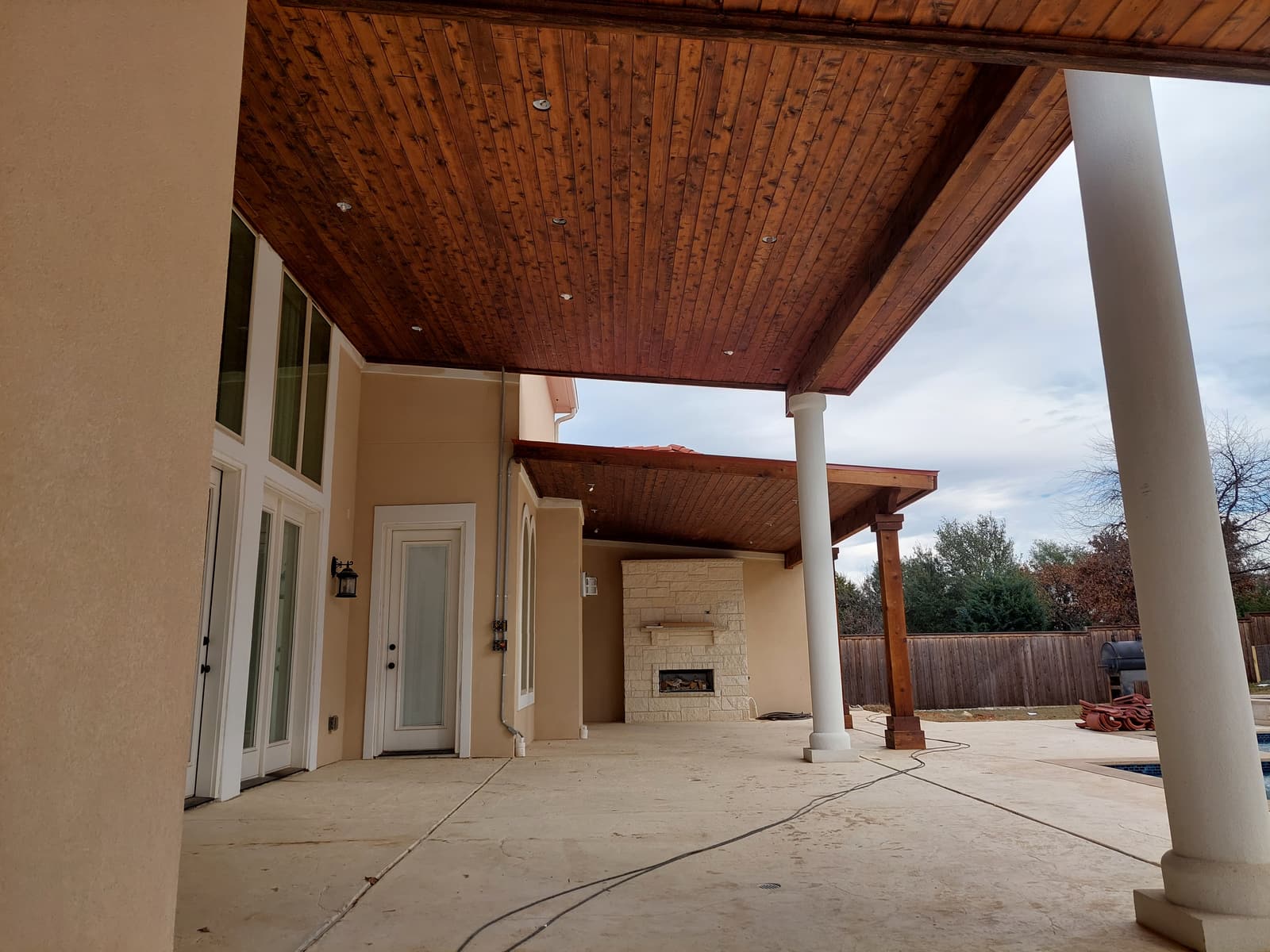 Covered patio with a stained wood ceiling, white columns and a stone outdoor fireplace with a raised hearth.