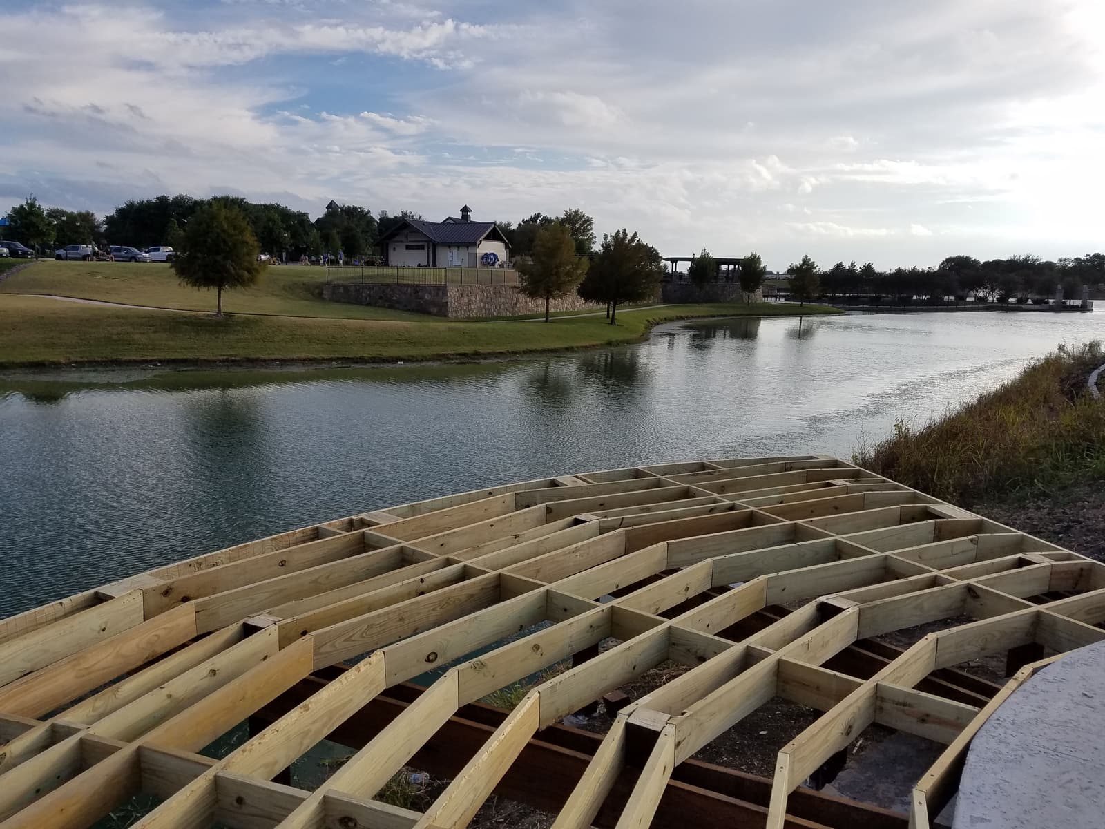 Treated lumber joist framing for a curved dock deck, laid out over a pond with a park pavilion visible across the water.