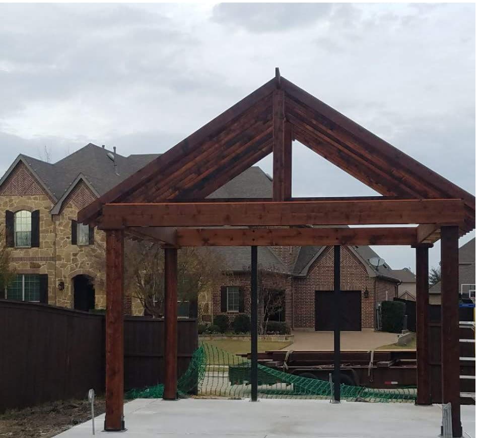 Freestanding stained timber pavilion frame with a steep gable and exposed king-post truss, standing on a new concrete patio behind a stone-and-brick home.