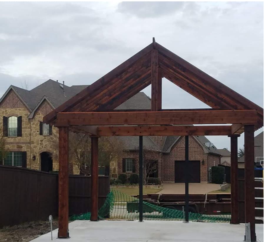Freestanding stained timber pavilion frame with a steep gable and exposed king-post truss, standing on a new concrete patio behind a stone-and-brick home.