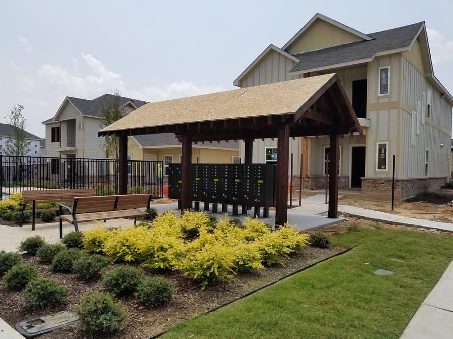 Timber-frame mail kiosk pavilion with stained posts and a gable roof, sheltering cluster mailboxes at a residential community.