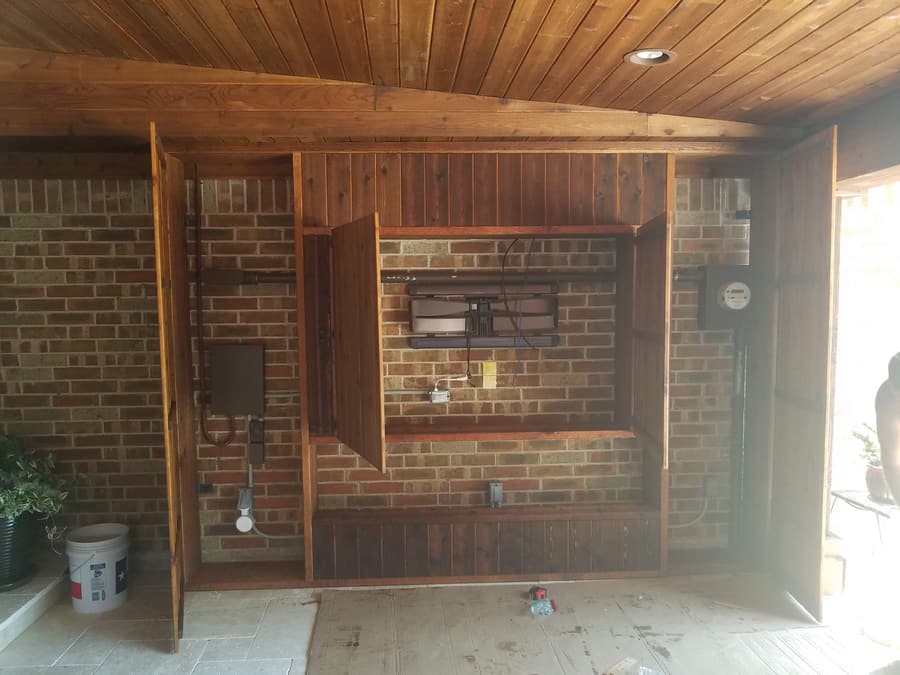 Covered patio under construction showing a cedar media wall framed over brick with an articulating TV mount, gas line and electrical rough-in exposed.