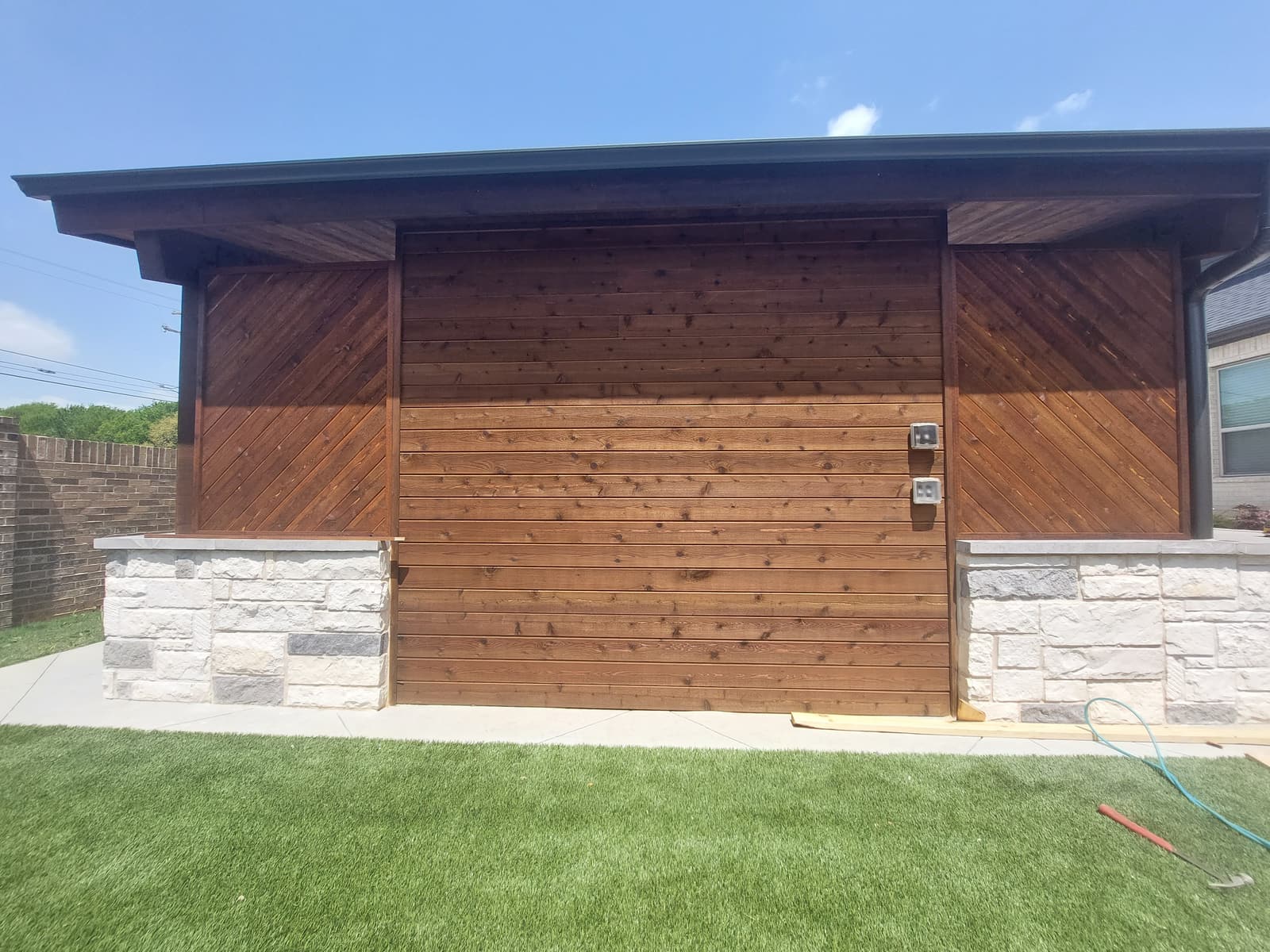 Modern shed-roof cabana with horizontal and chevron-pattern stained cedar siding above a white stone base, set on artificial turf.