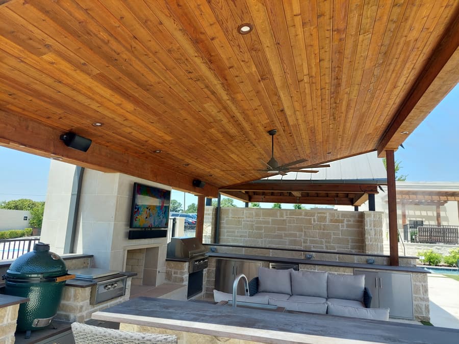 Poolside outdoor kitchen under a stained cedar tongue-and-groove pavilion ceiling, with a built-in grill, Big Green Egg, stone fireplace and mounted television.