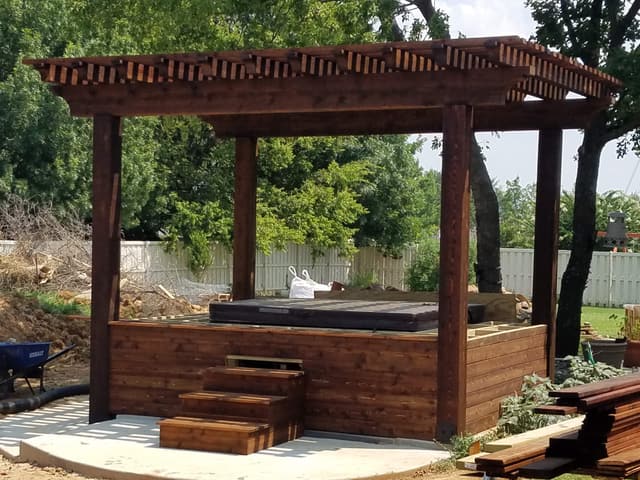 Freestanding dark-stained pergola over a hot tub, with a cedar skirt enclosing the spa and matching steps leading up to it.