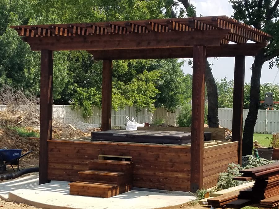 Freestanding dark-stained pergola over a hot tub, with a cedar skirt enclosing the spa and matching steps leading up to it.