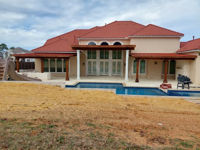Two matching stained cedar pergolas attached to either side of a large stucco home with a red tile roof, framing a swimming pool.
